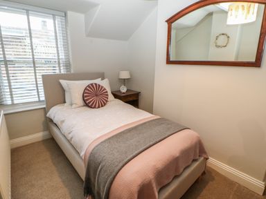 A bedroom with a single bed with pink and gray blankets a round pillow a wooden nightstand with a lamp and clock a window with blinds and a wall mirror at Chapel Cottage in Corbridge