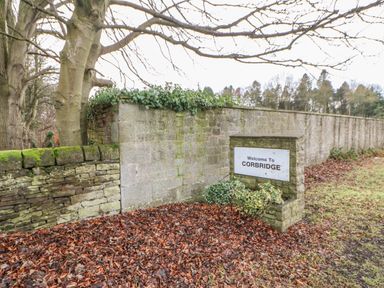 A stone wall with a welcome to Corbridge sign near trees and fallen leaves at Chapel Cottage in Corbridge