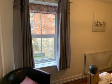 A room with a window showing a brick wall outside gray curtains a black chair with a cushion and a radiator with a painting of birds on the wall at A Stone's Throw in Alnmouth