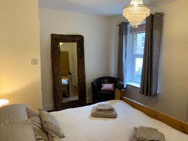 A bedroom with a wooden bed, pillows, folded towels, a large wooden framed mirror, black armchair, window with curtains and a chandelier at A Stone's Throw in Alnmouth