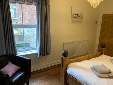 A bedroom with a wooden bed, a black armchair with a pink pillow, a window with curtains, and a door at A Stone's Throw in Alnmouth