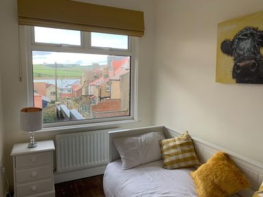 A bedroom with a white bed with cushions next to a window with a view of rooftops and hills at A Stone's Throw in Alnmouth