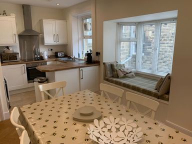 A kitchen with white cabinets a wooden countertop a dining table with bee patterned tablecloth and a window seat with cushions at A Stone's Throw in Alnmouth