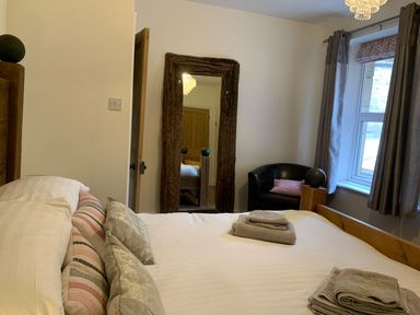 A bedroom with a bed pillows towels a wooden framed mirror a black chair and curtains at A Stone's Throw in Alnmouth