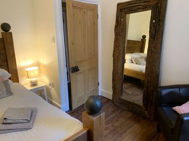 A bedroom with a wooden bed with towels on it a bedside table with lamp a wooden door a large floor mirror with wooden frame and a black armchair at A Stone's Throw in Alnmouth