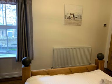 A bedroom with a wooden bed frame a window with curtains a radiator and a painting of birds on the wall at A Stone's Throw in Alnmouth