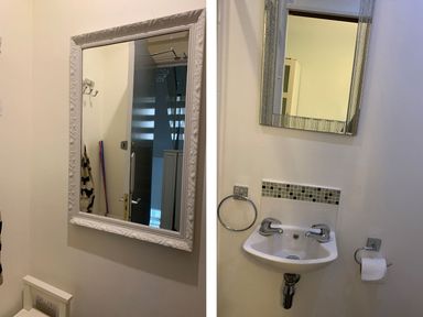 A wall mirror above a small white sink with two taps and a tiled backsplash in a bathroom with a towel ring and toilet paper holder