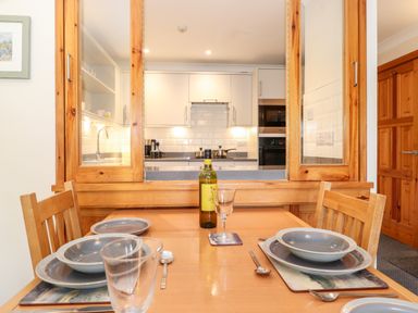 A dining table set with plates bowls glasses and a bottle of wine next to a kitchen with white cabinets and wooden framed pass through window at Pavey Ark in Ambleside