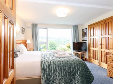 A bedroom with a bed covered by a quilt and towels placed on it a TV on a stand wooden furniture and a window with curtains at Pavey Ark Ambleside