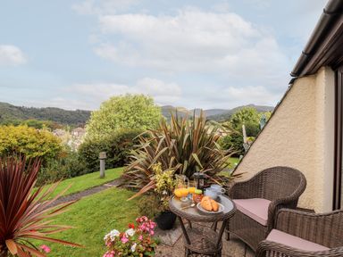A small outdoor patio area with two wicker chairs and a round table set with croissants orange juice and mugs in a garden with bushes and distant hills at Pavey Ark in Ambleside