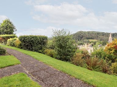 A garden path surrounded by trimmed bushes and green grass with hills and a church spire in the background at Pavey Ark in Ambleside