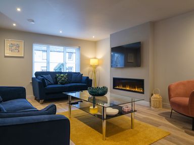 A living room with blue sofas a glass coffee table a yellow rug a wall-mounted television and a fireplace at The Neon Rose in Bowness-On-Windermere