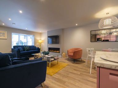 A living room with blue sofas a glass coffee table a wall-mounted TV an electric fireplace a round pink chair and a dining area with white chairs and a marble countertop at The Neon Rose in Bowness-On-Windermere