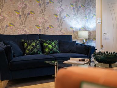 A living room with a dark blue sofa and patterned cushions a glass coffee table with a book and decorative bowl a table lamp and floral bird wallpaper at The Neon Rose in Bowness-On-Windermere