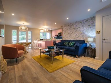 An open plan living room and kitchen with blue sofas a glass coffee table on a yellow rug a dining table with chairs and a pink kitchen at The Neon Rose in Bowness-On-Windermere