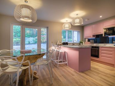 A kitchen with pink cabinets a marble countertop a glass table with white chairs and pendant lights at The Neon Rose in Bowness-On-Windermere