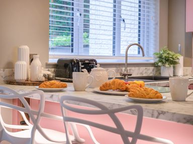 A kitchen counter with croissants on plates mugs and a teapot near a window with blinds at The Neon Rose in Bowness-On-Windermere