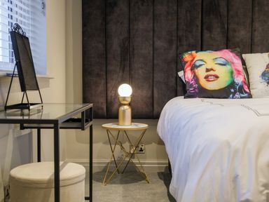 A bedroom corner with a bed with colorful face pillow a small round table with a lamp and a glass desk with a stool at The Neon Rose in Bowness-On-Windermere