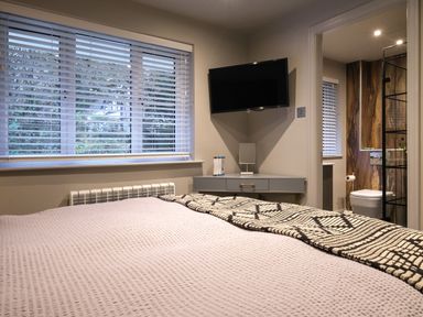 A bedroom with a bed a window with blinds a wall mounted television and an en suite bathroom at The Neon Rose in Bowness-On-Windermere