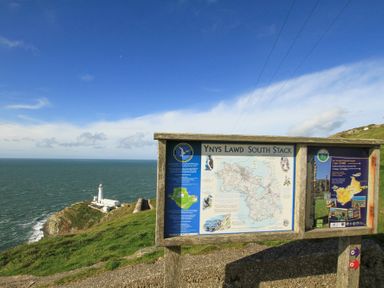 An outdoor information board with maps and text overlooking a lighthouse on a cliff by the sea at Glasfryn in Holyhead