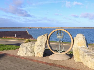 A metal anchor with a crown inside a circular stone sculpture near a waterfront with boats and a pier in the background at Glasfryn in Holyhead