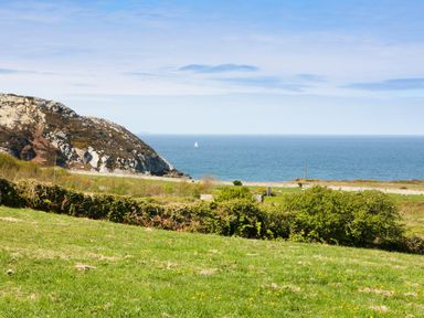 A coastal landscape with grassy fields bushes rocky hill and a sailboat in the sea at Glasfryn in Holyhead