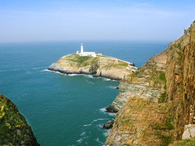 A lighthouse on a rocky island surrounded by ocean with cliffs in the foreground at Glasfryn in Holyhead