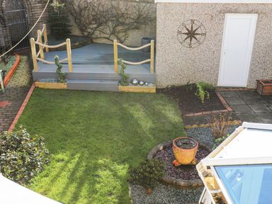 A backyard garden with a small lawn a deck with rope railing a white door a compass wall decor and potted plants at Glasfryn in Holyhead