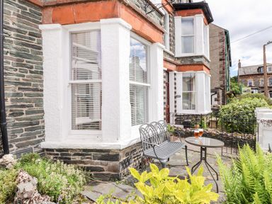 A small outdoor patio with a metal bench and table holding two glasses in front of a stone house with white framed windows at Oakhurst in Keswick