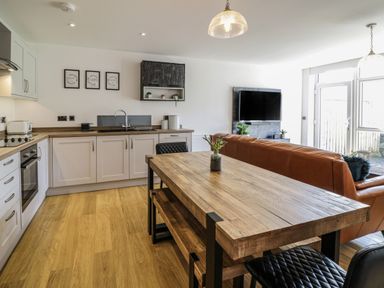 An open plan kitchen and living area with a wooden dining table black chairs a brown sofa a wall-mounted TV and kitchen cabinets at 2 Pensyflog Barns in Porthmadog