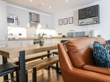 An open kitchen and dining area with wooden table, black chairs, white cabinets, and a brown leather sofa with cushions at 2 Pensyflog Barns in Porthmadog