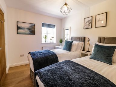 A bedroom with two single beds with blue throws and pillows a window with a striped blind and framed pictures on the wall at 2 Pensyflog Barns in Porthmadog
