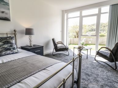 A bedroom with a metal bed frame bedside table lamp two chairs and large windows overlooking a garden at 2 Pensyflog Barns in Porthmadog