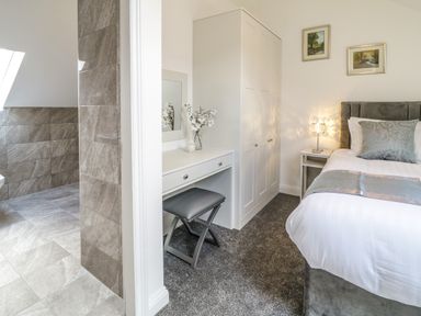 A bedroom with a bed a side table with a lamp a wardrobe a dressing table and a stool next to a bathroom with tiled walls and floor at 2 Pensyflog Barns Porthmadog