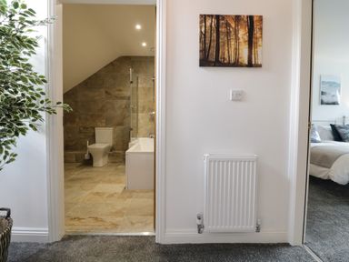 A hallway with a radiator and a picture on the wall leading to a bathroom with a toilet and bathtub and a bedroom with a bed at 2 Pensyflog Barns in Porthmadog