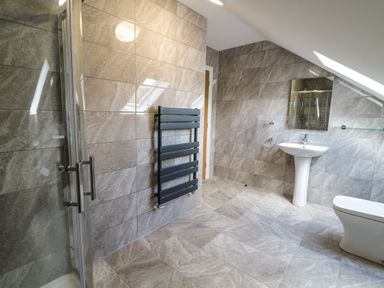 A bathroom with tiled walls and floor a shower a black towel radiator a pedestal sink a mirror and a toilet at 2 Pensyflog Barns in Porthmadog