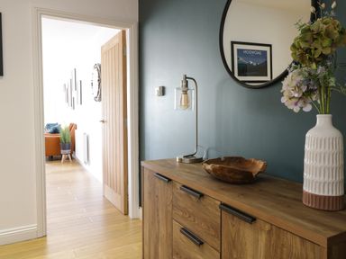 A wooden sideboard with a lamp a wooden bowl and a vase with flowers next to a teal wall and doorway at 2 Pensyflog Barns in Porthmadog