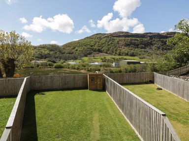 A fenced garden with a wooden shed and green grass with trees and hills in the background at 2 Pensyflog Barns in Porthmadog