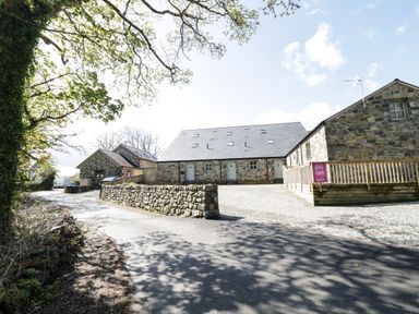 A stone building with a paved driveway and trees in the background at 2 Pensyflog Barns in Porthmadog