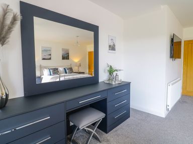 A bedroom with a dark blue dresser mirror and stool with a bed reflected in the mirror at 2 Pensyflog Barns Porthmadog