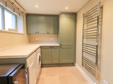 A utility room with green cabinets a washing machine a dryer a countertop a window and a heated towel rail at Old Howgate Barn Outgate near Hawkshead