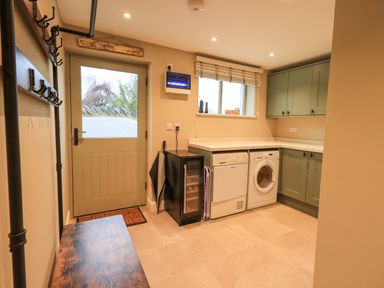 A utility room with a bench and coat hooks on the left a door a small fridge dishwasher washing machine green cabinets and a window at Old Howgate Barn Outgate near Hawkshead