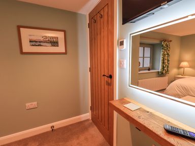 A bedroom corner with a wooden door a wall painting a mirror with lights a remote on a wooden shelf and a window with green patterned curtains at Old Howgate Barn Outgate near Hawkshead