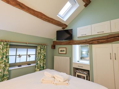 A bedroom with a bed with folded towels a window with green leaf patterned curtains a wall mounted TV and white cupboards at Old Howgate Barn Outgate near Hawkshead