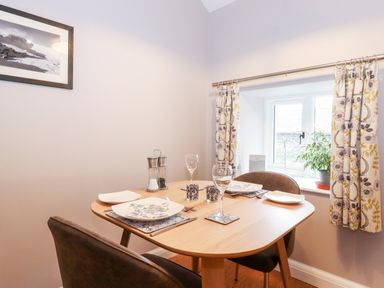 A dining area with a wooden table set for two near a window with patterned curtains and a potted plant at Low Ashmead Cottage in Hawkshead