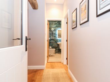 A hallway with wooden floor and framed pictures on the wall leading to a bathroom with a toilet and sink at Low Ashmead Cottage in Hawkshead