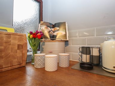 A kitchen countertop with a wooden cutting board, a jar of tulips, ceramic canisters, a bread bin with an open cookbook, stacked mugs in a rack, and an electric kettle at Rayside Ullswater
