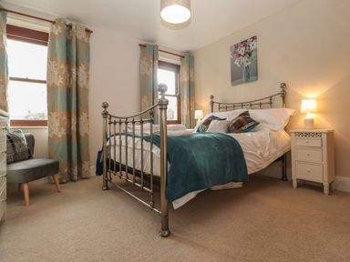 A bedroom with a metal bed frame a teal blanket two windows with curtains bedside table lamps and a gray chair at Rayside in Ullswater
