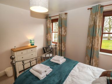 A bedroom with a metal bed frame towels on bed a chest of drawers with a lamp a chair and floral curtains at Rayside in Ullswater