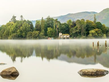 A lake with rocks in the foreground and a small house surrounded by trees with hills in the background at Rayside in Ullswater